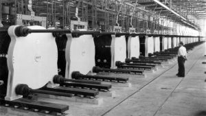 Black and white photo of electolyser in the 1950s. A plant worker stand in front of the machine that stretches the length of a large warehouse