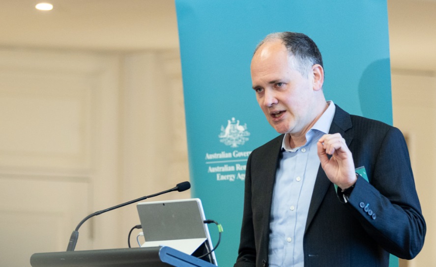 Alex Campbell stands behind a podium with his left hand raised, talking to an ARENA Insights forum audience